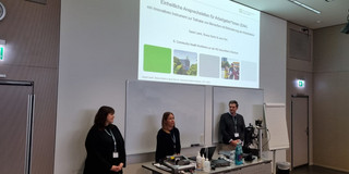 Teresa Sartor, Dr. Jana York und Daniel Krüger (v.l.n.r.) bei ihrem Vortrag über Einheitliche Ansprechstellen für Arbeitgeber*innen. Sie stehen hinter einem grauen Metalltisch mit dem Rücken zu einer weißen Wand, auf der die Projektion der ersten Folie ihres Vortrags mit dessen Titel zu sehen ist.